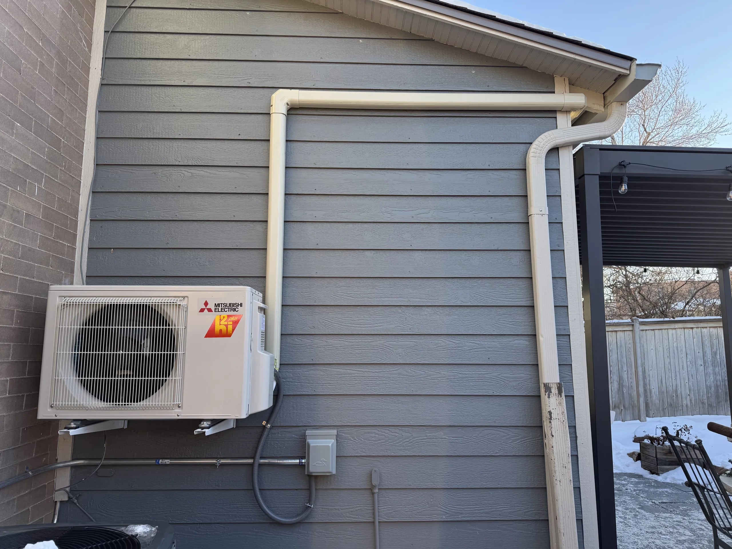 A compact Mitsubishi heat pump mounted on blue-gray siding, with snow in the yard